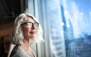 Vanhempi silmälasipäinen ja hymyilevä nainen katsoo ulos sinisestä ikkunasta, johon heijastuu ulkona näkyviä rakennuksia.