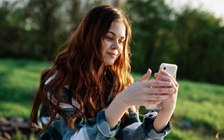 Kuvassa nuori nainen katsoo käsissään olevaa valkoisen kännykän näyttöä ja hymyilee. Naisella on pitkät ruskeat hiukset ja sinivalkoinen ruutupaita. Taustalla näkyy vihreää luontoa.